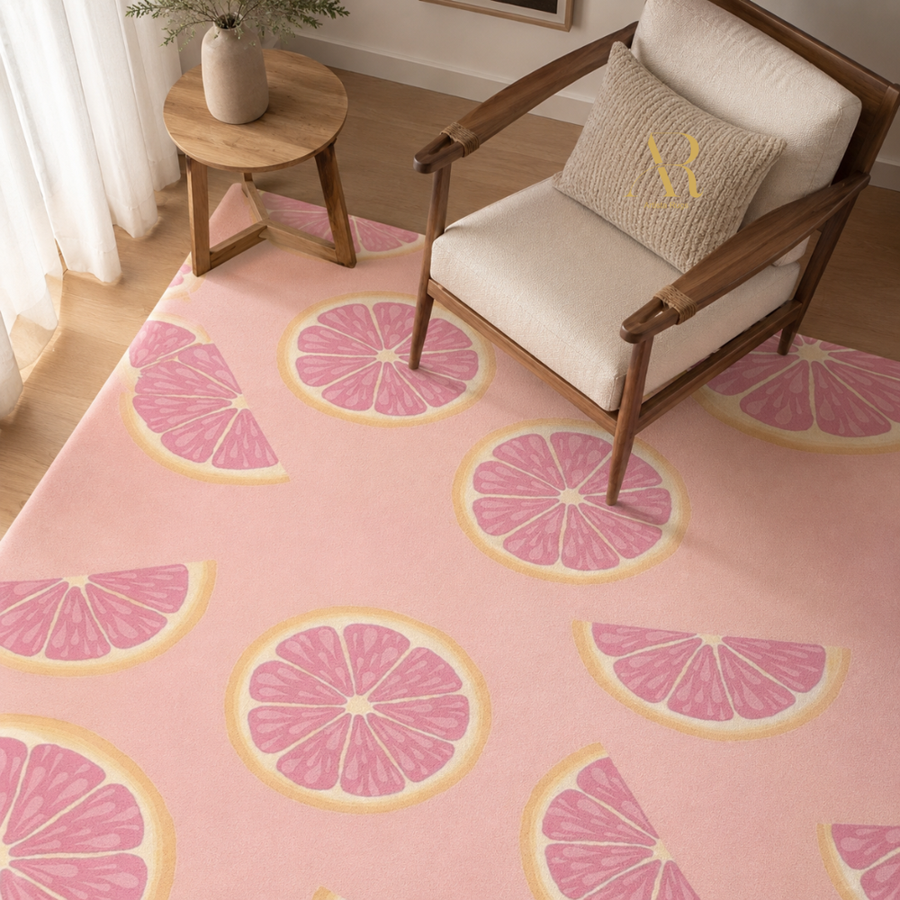 A pink area rug with a repeating pattern of pink grapefruit slices, styled in a bright room with a wooden armchair, a side table, and a decorative pillow.