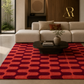 A modern living room scene featuring a large area rug with a bold geometric pattern in red and dark burgundy. The rug is styled in a contemporary interior with a cream sectional sofa, a dark wood coffee table, and a vase with branches.