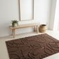 Brown textured rug on a light wooden floor with a bench and plant in the background.