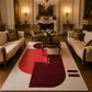 A large modern area rug with an abstract geometric pattern in cream, red, pink, and burgundy, styled in a luxurious, symmetrical vintage living room with cream tufted sofas and a dark wood coffee table.