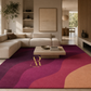 A large area rug with a wavy abstract pattern in magenta, burgundy, pink, and peach tones, styled in a modern living room with a beige sectional sofa and a wooden coffee table. The brand name 'Arts & Roes' with a gold monogram logo is printed on the rug.