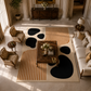 A styled living room scene with a large area rug as the focal product. The rug has a cream base with bold black organic shapes and warm tan vertical stripes.