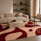 A modern living room scene featuring a large area rug with an abstract organic pattern in deep red and beige. The rug is styled in a contemporary living space with a cream-colored sectional sofa, a stone coffee table, a wooden armchair, and built-in floating shelves in the background.