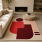 A modern abstract area rug with geometric shapes in red, pink, and dark maroon on a cream base, styled in a contemporary living room next to a beige sofa and a round stone coffee table.
