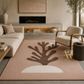 A large rectangular area rug in a modern living room setting. The rug has a dusty pink base with a large dark brown abstract tree or coral-like motif and a small off-white semicircle at the bottom.