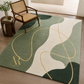 A rectangular area rug with an abstract wavy pattern in sage green, cream, and dark forest green, accented by flowing gold lines. The rug is styled in a modern living room setting on a light wood floor next to a mid-century modern armchair and a small wooden side table.