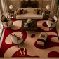 A luxury living room scene featuring a large abstract patterned area rug in deep red and beige. The rug is styled in an elegant room with a cream-colored sofa, matching armchairs, a dark wood coffee table, and a tufted bench.