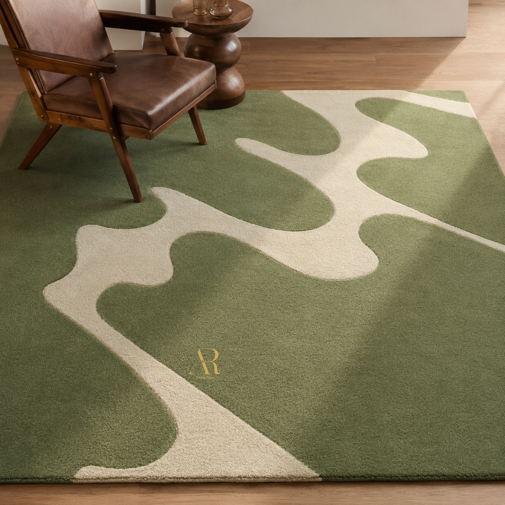 A green area rug with an abstract wavy pattern in a light beige color, styled in a room with a wooden floor, a mid-century modern armchair, and a small wooden side table. A gold monogram letter 'R' is visible on the rug.