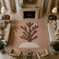 A large rectangular area rug in a styled living room. The rug has a dusty pink background with a large dark brown coral-like tree motif in the center, a cream arch beneath it, and a gold monogram logo reading 'Artema Realty' on the left.