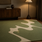 A green area rug with a cream wavy abstract pattern and a small gold logo, styled in a modern living room setting next to a wooden sideboard and a floor lamp.