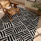 A black and off-white area rug with a bold abstract brushstroke pattern, styled in a modern living room setting with a wooden armchair and a potted plant.