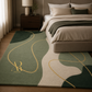 A modern area rug with an abstract organic pattern in sage green, cream, and dark forest green, accented with flowing gold lines. The rug is styled in a bedroom setting next to a bed with neutral bedding.