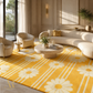 A bright yellow area rug with a white daisy and stripe pattern in a modern, sunlit living room with cream-colored furniture.