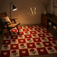 A rectangular area rug with a checkerboard pattern of red squares, cream squares, red roses, and cream hearts, styled in a warm, dimly lit living room next to a wooden armchair and a floor lamp.