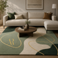 A modern living room scene featuring a large area rug with abstract wavy patterns in sage green, cream, and dark forest green, accented with gold lines. The rug is styled in a living room setting with a beige sectional sofa, a round wooden coffee table, and neutral-toned decor.