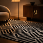 A black and off-white geometric patterned area rug styled in a warm, modern living room setting next to a wooden cabinet and a floor lamp.
