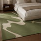 A green area rug with an abstract wavy pattern in cream, shown on a dark wood floor in a modern bedroom setting next to a bed.