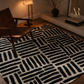 A modern area rug with a bold black and off-white geometric pattern, styled in a warm, contemporary living room setting next to a wooden armchair.
