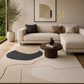 A modern living room scene featuring a large abstract-patterned area rug in beige, cream, and charcoal gray. The rug is styled in a contemporary living room with a light beige sectional sofa, a round dark wood coffee table, and a potted olive tree by a window.