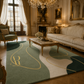 An elegant living room featuring a large area rug with abstract green, cream, and dark teal shapes outlined in gold. The rug is styled in a luxurious room with a matching green armchair, a cream sofa, a marble-top coffee table, and ornate gold-framed furniture.