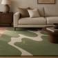 A green and beige abstract-patterned area rug styled in a modern living room setting with a beige sofa, wooden side table, and a lamp in the background.