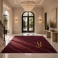 A plush burgundy area rug with elegant wavy gold lines and a gold monogram logo, displayed in a luxurious, sunlit entryway with marble floors and a crystal chandelier.