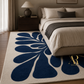 A large area rug with a bold navy blue abstract floral pattern on a cream background, styled in a modern bedroom next to a bed.