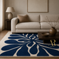 A large area rug with a bold navy blue and cream floral pattern, styled in a modern living room setting with a beige sofa, wooden coffee table, and neutral decor.