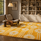 A large yellow area rug with a white floral and stripe pattern is shown in a styled living room setting with a wooden lounge chair, floor lamp, and built-in bookshelves.