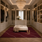 A luxury walk-in closet interior with built-in cabinetry, a large crystal chandelier, a white bench, and a deep burgundy area rug featuring a gold monogram logo. Clothes are displayed on racks and shelves, and the room is lit by warm ambient lighting.