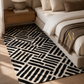 A modern area rug with a bold black and off-white geometric pattern styled in a contemporary bedroom next to a bed with neutral-toned bedding and a wooden nightstand.