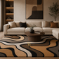 A modern living room featuring a large area rug with a bold, wavy abstract pattern in beige, brown, black, and cream. The room is styled with a cream-colored sectional sofa, a round wooden coffee table, and warm ambient lighting.