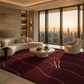 A luxury modern living room featuring a large deep burgundy area rug with wavy white lines and a gold 'Alessio Rognoni' monogram. The room is styled with a curved cream sectional sofa, a matching accent chair, and a round marble coffee table, set against a backdrop of floor-to-ceiling windows showing a city skyline at sunset.