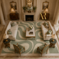 An overhead view of an elegant living room featuring a large light green and cream wavy patterned area rug. The room is furnished with two cream sofas, two matching armchairs, two small marble-top side tables with lamps, a central coffee table with a floral arrangement, and a fireplace in the background.