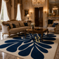 A large cream-colored area rug with a bold navy blue floral or leaf-like pattern, styled in an elegant, luxurious vintage living room with gold furniture and chandeliers.