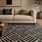 A modern black and beige geometric patterned area rug in a styled living room setting with a beige sofa, decorative pillows, and a wooden coffee table.