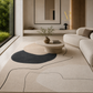 A large modern area rug with abstract organic shapes in beige, cream, and charcoal gray, styled in a minimalist, warm-toned living room with a curved sofa and a low stone coffee table.