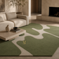 A green and beige abstract patterned area rug styled in a modern, warm-toned living room with a cream sofa, stone coffee table, and fireplace.
