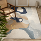 An abstract area rug with a modern design in beige, navy, light blue, and gray, styled in a bright room next to a wooden armchair and a potted plant.