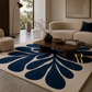 A large cream-colored area rug with a bold navy blue floral or fan-like pattern, styled in a modern living room setting with a wooden coffee table, beige sofa, and neutral decor.
