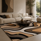 A modern living room scene featuring a large area rug with a wavy, abstract pattern in beige, brown, black, and cream. The rug is styled in a contemporary living room with a cream sectional sofa, a round dark coffee table, and a potted plant.