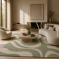 A modern living room featuring a large area rug with a wavy abstract pattern in sage green and cream. The room is styled with a curved beige sofa, a matching armchair, a low stone coffee table, and warm neutral walls.