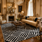 A luxury living room featuring a large black and white geometric patterned area rug on a parquet floor, styled with an elegant cream sofa, gold accents, and ornate chandeliers.