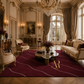 An ornate luxury living room with cream upholstery furniture, a large deep burgundy area rug with wavy white lines and a gold monogram logo, and a crystal chandelier.