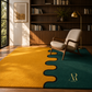 A modern area rug in a warm yellow and deep teal green color-blocked design with a wavy black dividing line, styled in a sunlit room with a mid-century armchair and wooden bookshelves.
