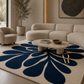 A large modern area rug in a styled living room, featuring a bold navy blue floral or leaf-like pattern on a warm cream background. The rug is styled with a round stone coffee table, a curved cream boucle sofa, and a matching armchair.