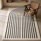 A rectangular area rug with a black arched stripe pattern on a cream background, styled in a modern room next to a wooden armchair.