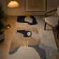 A rectangular area rug with an abstract organic pattern in beige, navy, blue, and gray, styled in a warm, modern living room next to a wooden armchair and a floor lamp.