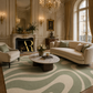 A luxury living room featuring a large sage green and cream wavy patterned area rug, a cream curved sofa, a matching armchair, and a round marble-top coffee table in an ornate classical room.