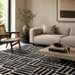 A modern living room scene featuring a black and off-white geometric patterned area rug, a light beige sofa, a mid-century wooden armchair, and a low concrete coffee table.